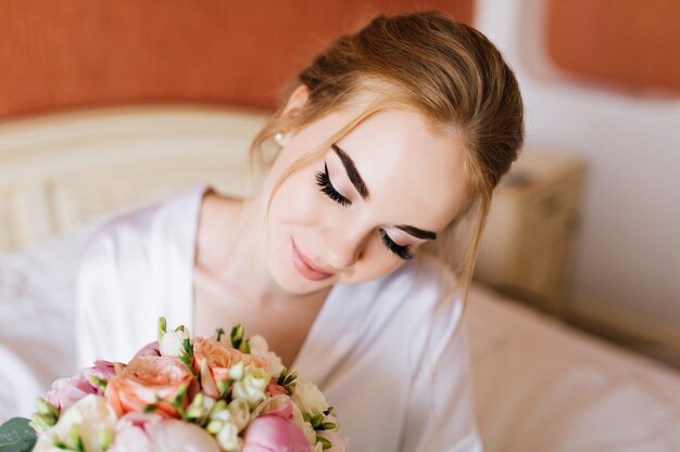 Closeup portrait jolie mariée heureuse en peignoir blanc le matin dans l'appartement. Elle regarde le bouquet de fleurs dans les mains et souriant