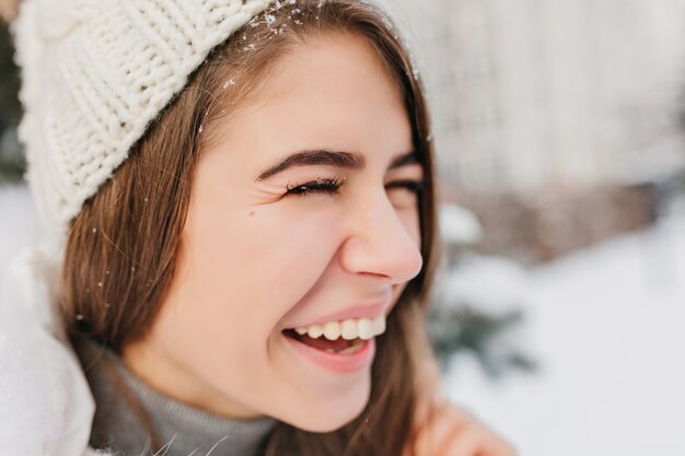 Closeup portrait lumineux visage positif émotions de femme joyeuse en bonnet tricoté blanc chaud en riant sur la rue pleine de neige. Vraies émotions, flocons de neige, s'amuser, profiter de l'hiver.