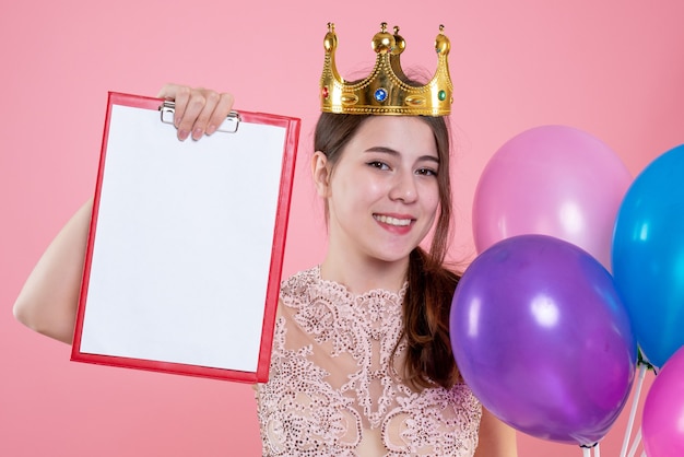 Closeup vue de face happy party girl avec couronne tenant le presse-papiers et les ballons