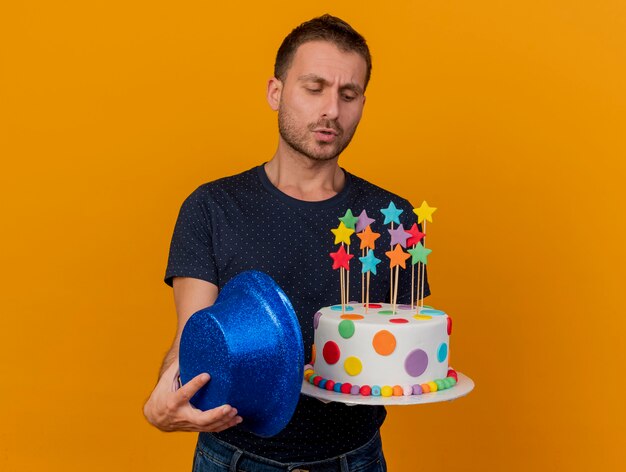 Clueless bel homme détient chapeau de fête bleu et regarde gâteau d'anniversaire isolé sur mur orange