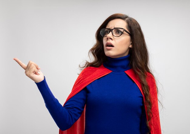 Clueless fille de super-héros caucasien avec cape rouge dans des lunettes optiques regarde et pointe sur le côté sur blanc