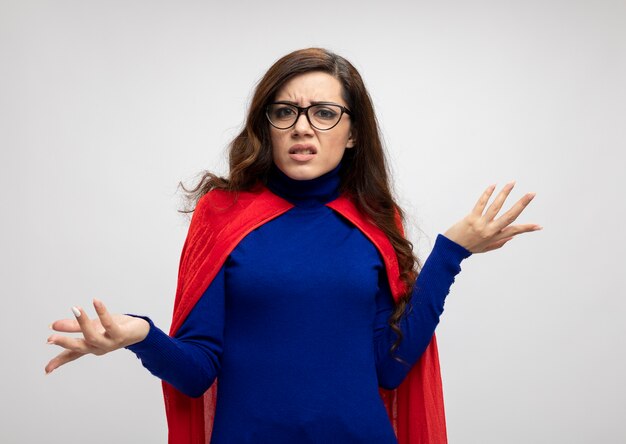 Clueless fille de super-héros caucasien dans des lunettes optiques avec cape rouge tient les mains ouvertes sur blanc