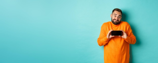 Photo gratuite clueless guy haussant les épaules et montrant l'émotion indécise de l'écran mobile debout dans un pull orange sur