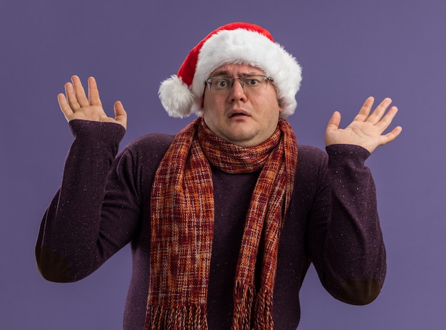 Photo gratuite clueless homme adulte portant des lunettes et bonnet de noel avec foulard autour du cou regardant la caméra montrant les mains vides isolés sur fond violet