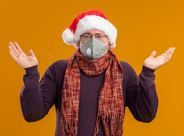 Clueless homme adulte portant des lunettes de masque de protection et bonnet de Noel avec foulard autour du cou regardant la caméra montrant les mains vides isolé sur fond orange