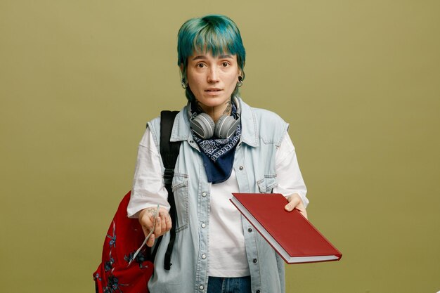 Clueless jeune étudiante portant des écouteurs et un bandana sur le cou et le sac à dos montrant de l'argent et un carnet de notes regardant la caméra isolée sur fond vert olive