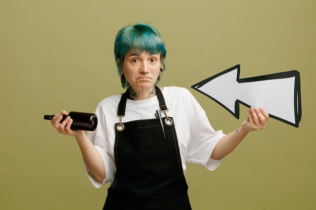 Clueless jeune femme barbier portant l'uniforme tenant le signe de la flèche pointant vers elle-même et la laque pour cheveux regardant la caméra isolée sur fond vert olive