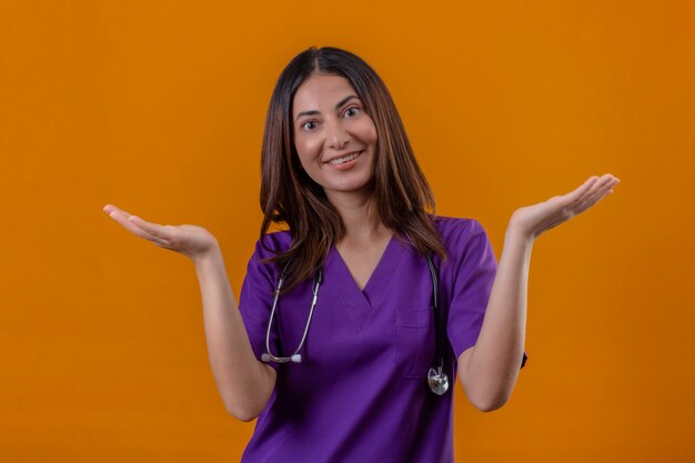 Clueless jeune femme infirmière en uniforme médical et avec stéthoscope en haussant les épaules à l'incertain et confus n'ayant pas de réponse étalant les paumes souriant debout sur orange isolé