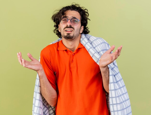 Clueless jeune homme malade portant des lunettes enveloppées dans un plaid à l'avant montrant les mains vides isolés sur mur vert olive