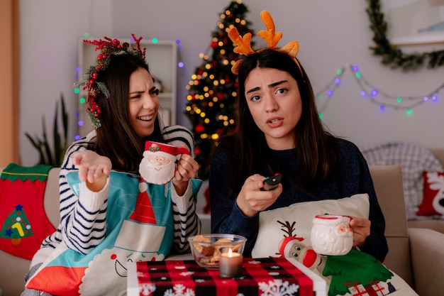 Clueless jolie jeune fille avec couronne de houx tient la tasse et regarde son ami tenant la télécommande du téléviseur assis sur un fauteuil le temps de Noël à la maison