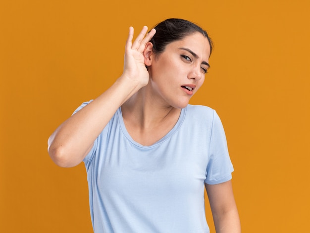 Clueless young brunette caucasian girl tient la main derrière l'oreille sur orange