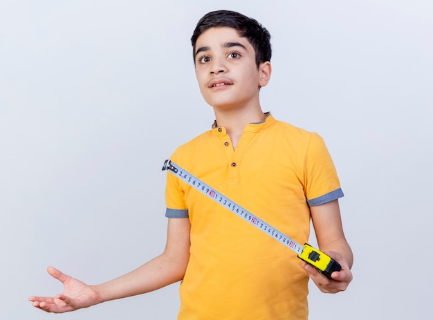 Clueless young caucasian boy holding tape mètre à droite montrant la main vide isolé sur fond blanc