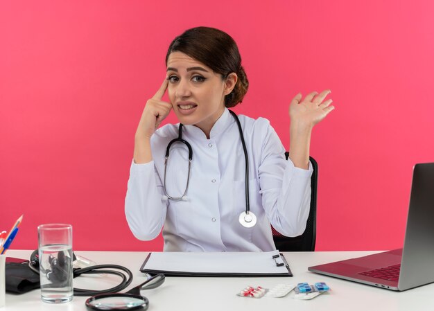 Clueless young female doctor wearing medical robe et stéthoscope assis au bureau avec des outils médicaux et un ordinateur portable pointant le doigt à la tête montrant la main vide isolée sur le mur rose