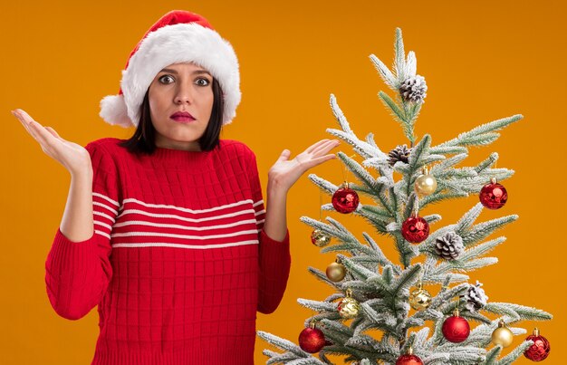 Photo gratuite clueless young girl wearing santa hat debout près de l'arbre de noël décoré montrant les mains vides isolés sur le mur orange