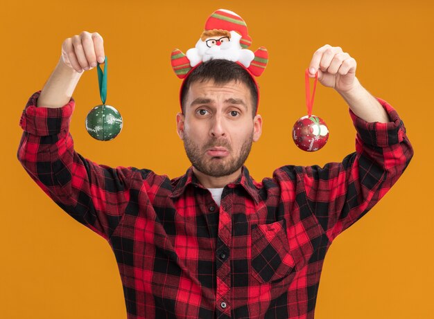 Clueless young man wearing santa claus headband regardant la caméra tenant des boules de Noël isolé sur fond orange