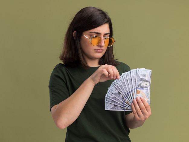 Clueless young pretty caucasian girl in sun lunettes tenant et regardant de l'argent sur vert olive