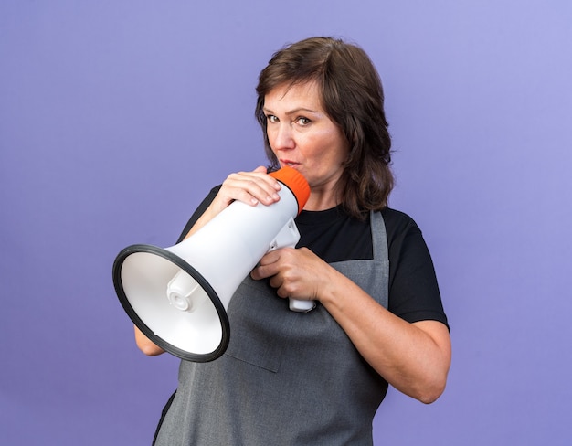 Coiffeur féminin de race blanche adulte impressionné en uniforme tenant un haut-parleur et regardant la caméra isolée sur fond violet avec espace de copie