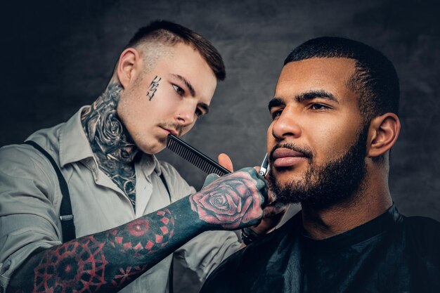 Coiffeur mâle caucasien tatoué coupant la barbe à l'homme élégant noir.