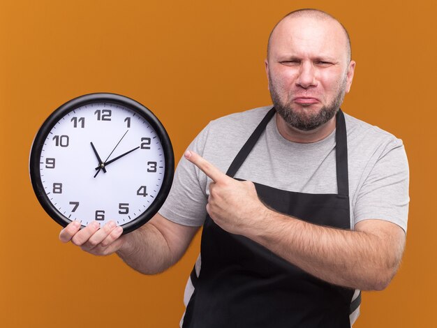 Un coiffeur masculin d'âge moyen mécontent en tenue uniforme et pointe sur une horloge murale isolée sur un mur orange