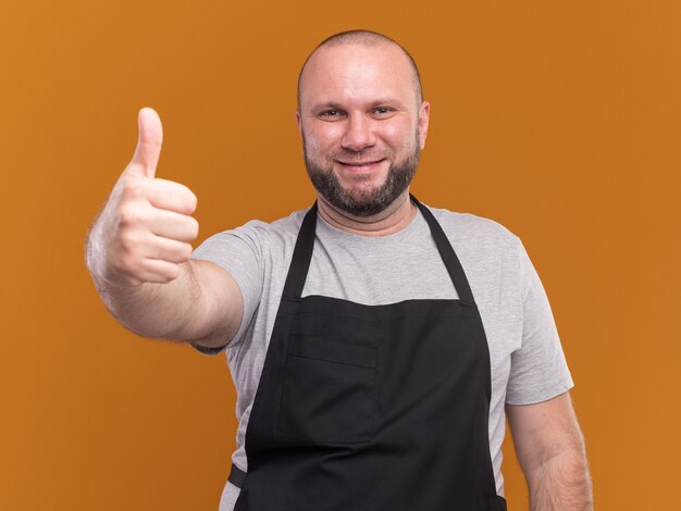 Coiffeur masculin d'âge moyen souriant en uniforme montrant le pouce vers le haut isolé sur un mur orange