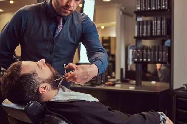 Coiffeur professionnel modelant la barbe avec des ciseaux et un peigne au salon de coiffure. Photo en gros plan.