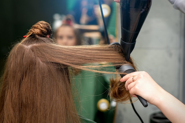 Un coiffeur sèche de longs cheveux bruns avec un sèche-cheveux et une brosse ronde dans un salon de coiffure.