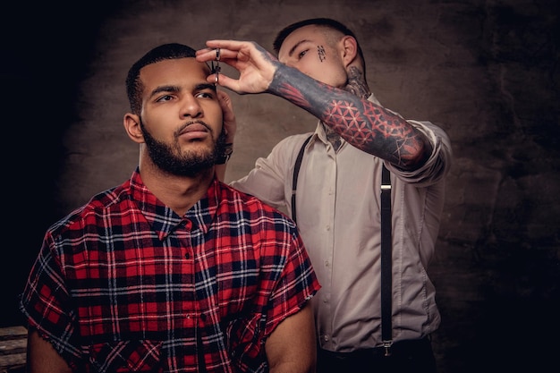 Photo gratuite un coiffeur tatoué professionnel à l'ancienne fait une coupe de cheveux à un client afro-américain, à l'aide de ciseaux et d'un peigne. isolé sur fond texturé sombre.