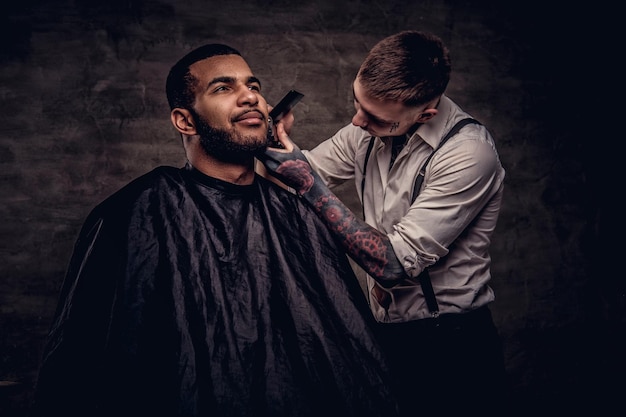 Photo gratuite un coiffeur tatoué professionnel à l'ancienne fait une coupe de cheveux à un client afro-américain, à l'aide d'une tondeuse et d'un peigne. isolé sur fond texturé sombre.