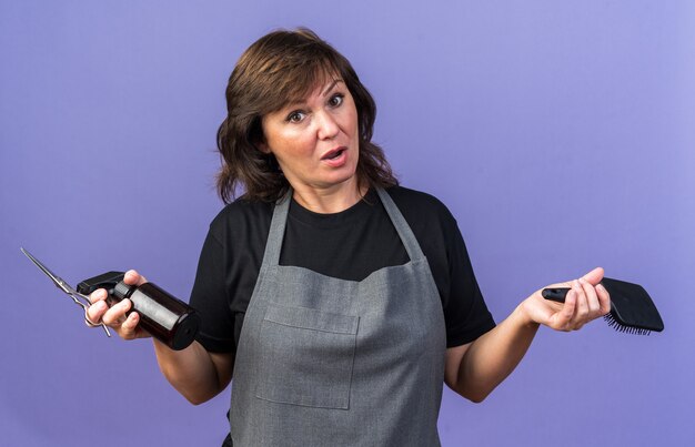 Coiffeuse caucasienne adulte surprise en uniforme tenant un peigne et des ciseaux pour flacon pulvérisateur isolés sur un mur violet avec espace pour copie