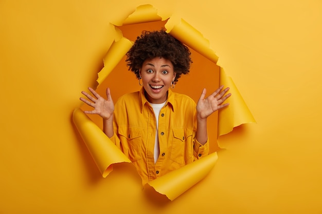 Photo gratuite la coiffure afro garde les paumes levées, profite de nouvelles agréables, a les cheveux bouclés naturels, montre l'excitation et le bonheur, se tient dans le trou déchiré du mur de papier jaune