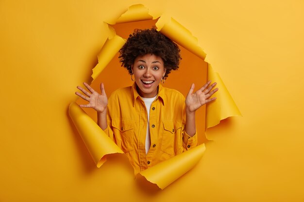 La coiffure afro garde les paumes levées, profite de nouvelles agréables, a les cheveux bouclés naturels, montre l'excitation et le bonheur, se tient dans le trou déchiré du mur de papier jaune