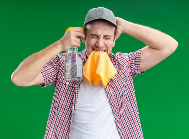 en colère contre les yeux fermés jeune homme nettoyeur portant une casquette tenant un chiffon de nettoyage sur la bouche montrant un geste de suicide avec un agent de nettoyage mettant la main sur la tête isolée sur un mur vert