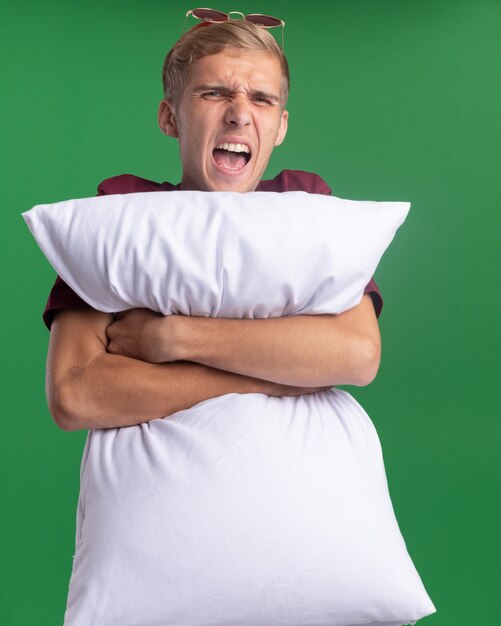 Colère jeune beau mec portant une chemise rouge oreiller étreint isolé sur mur vert