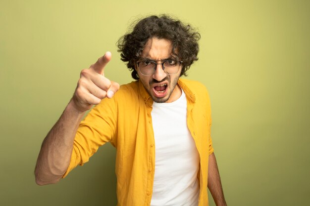 Colère jeune bel homme portant des lunettes à la recherche et pointant vers l'avant isolé sur mur vert olive