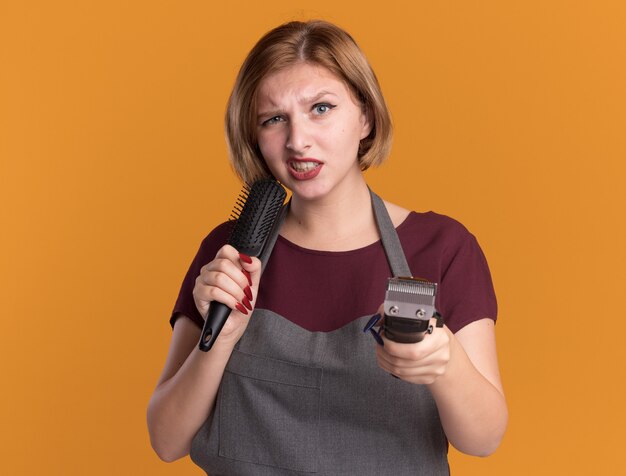 En colère, jeune, belle femme, coiffeur, dans, tablier, tenue, tondeuse, et, cheveux, brushlooking, devant, mécontent, debout, sur, mur orange