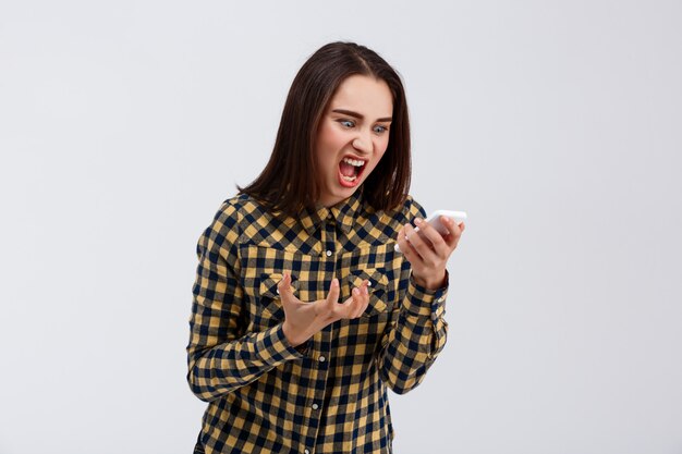 En colère jeune belle fille vêtue d'une chemise à carreaux criant au téléphone sur le mur blanc.