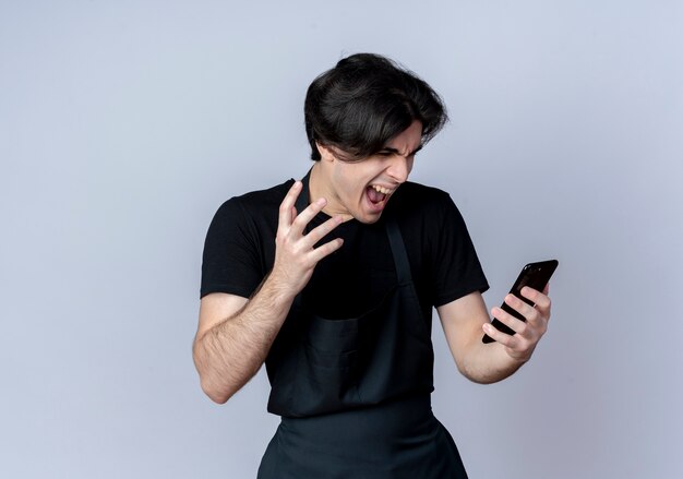 En colère jeune homme beau coiffeur en uniforme tenant et regardant le téléphone isolé sur fond blanc