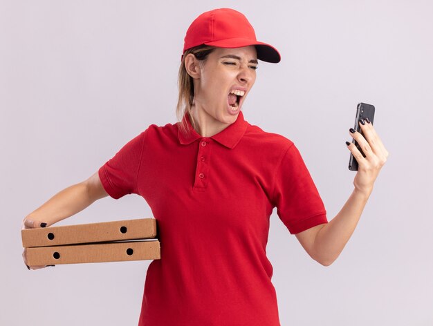 Colère jeune jolie livreuse en uniforme détient des boîtes à pizza et regarde le téléphone sur blanc