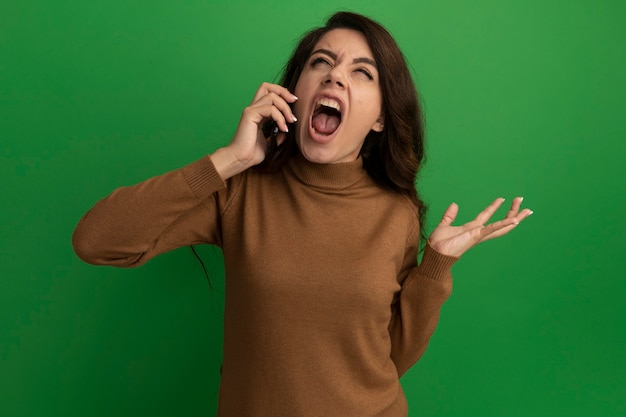 En colère en levant la belle jeune fille parle au téléphone et répandant la main isolée sur le mur vert