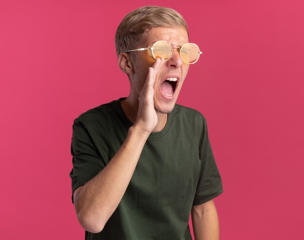 En colère regardant côté jeune beau mec portant une chemise verte et des lunettes appelant quelqu'un isolé sur un mur rose