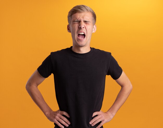 En colère avec les yeux fermés jeune beau mec vêtu d'une chemise noire mettant les mains sur la hanche isolé sur mur jaune