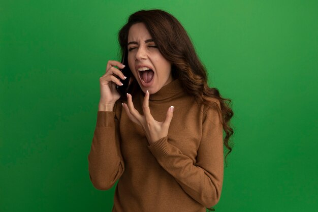 En colère, les yeux fermés, jeune belle fille parle au téléphone isolé sur un mur vert