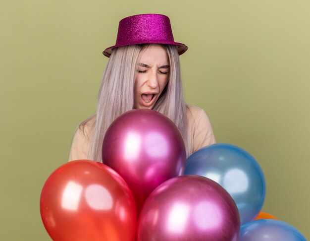 En colère avec les yeux fermés jeune belle fille portant un chapeau de fête debout derrière des ballons