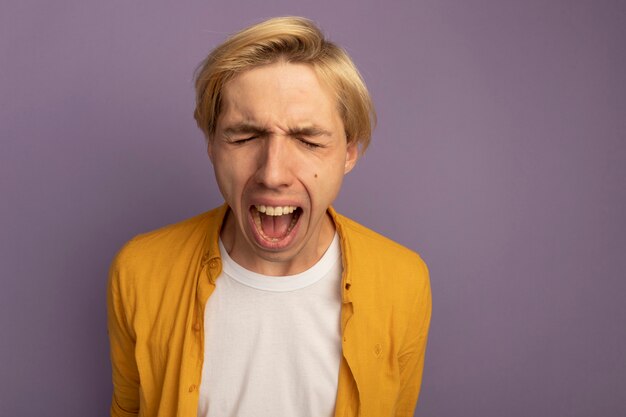 En colère, les yeux fermés, jeune mec blond portant un t-shirt jaune isolé sur violet avec espace copie