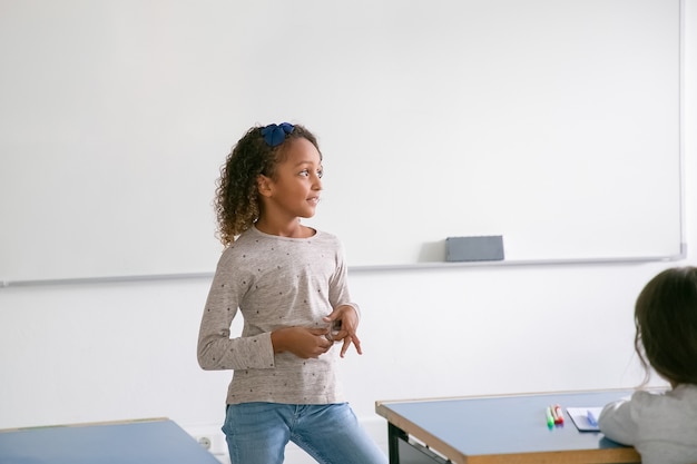 Écolière afro-américaine souriante pensif debout au tableau blanc en face de la classe