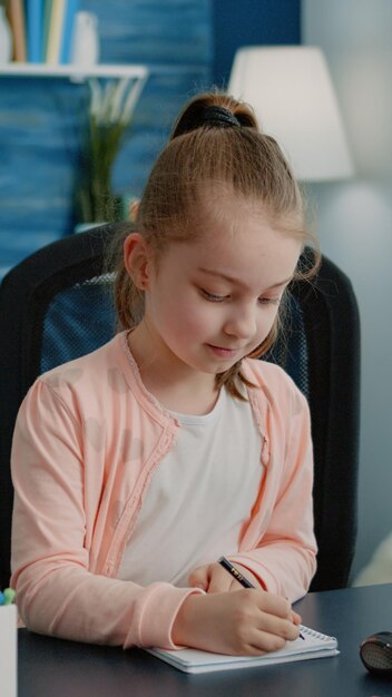 Écolière écrivant sur un cahier avec un stylo pour les devoirs. Le petit enfant s'est concentré sur la tâche pour les cours à distance en ligne tout en étant assis au bureau avec un ordinateur et un manuel. Élève scolarisé à la maison