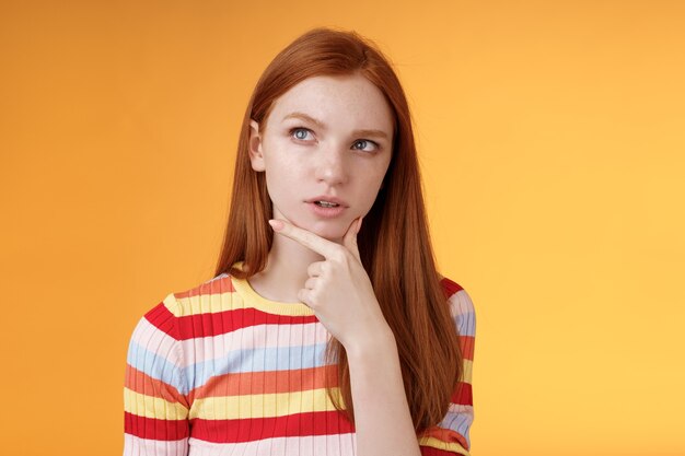 Une collègue féminine rousse à l'allure sérieuse et créative pensant à une mise au point profonde méditant sur une idée faisant un choix touchant le menton un regard réfléchi dans le coin supérieur gauche interrogé sur un fond orange perplexe.