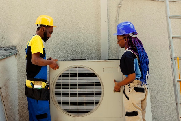 Des collègues ingénieurs afro-américains ouvrent un climatiseur rouillé hors service pour le remplacer par un nouveau condenseur extérieur performant après avoir vidé le réfrigérant et remplacé les conduits