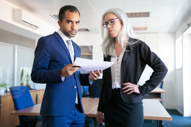 Collègues professionnels debout dans la salle de réunion avec des documents. Travailleuse aux cheveux gris focalisée dans le rapport de lecture de lunettes. Homme d'affaires regardant la caméra. Concept de travail d'équipe, d'entreprise et de gestion