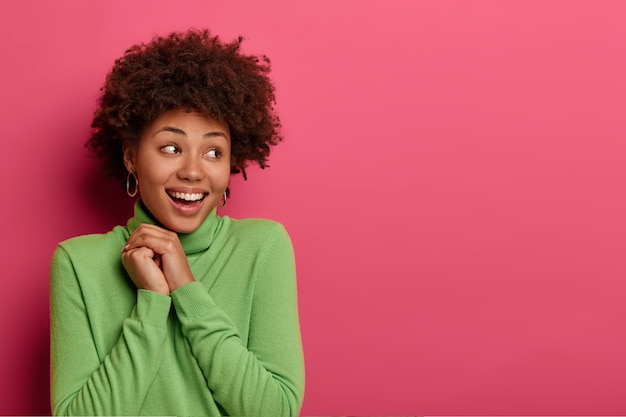 Photo gratuite comme c'est gentil! une jolie femme positive a les cheveux afro touffus, se détourne, admire quelque chose de très agréable, garde les mains jointes, sourit largement, a une peau sombre et saine